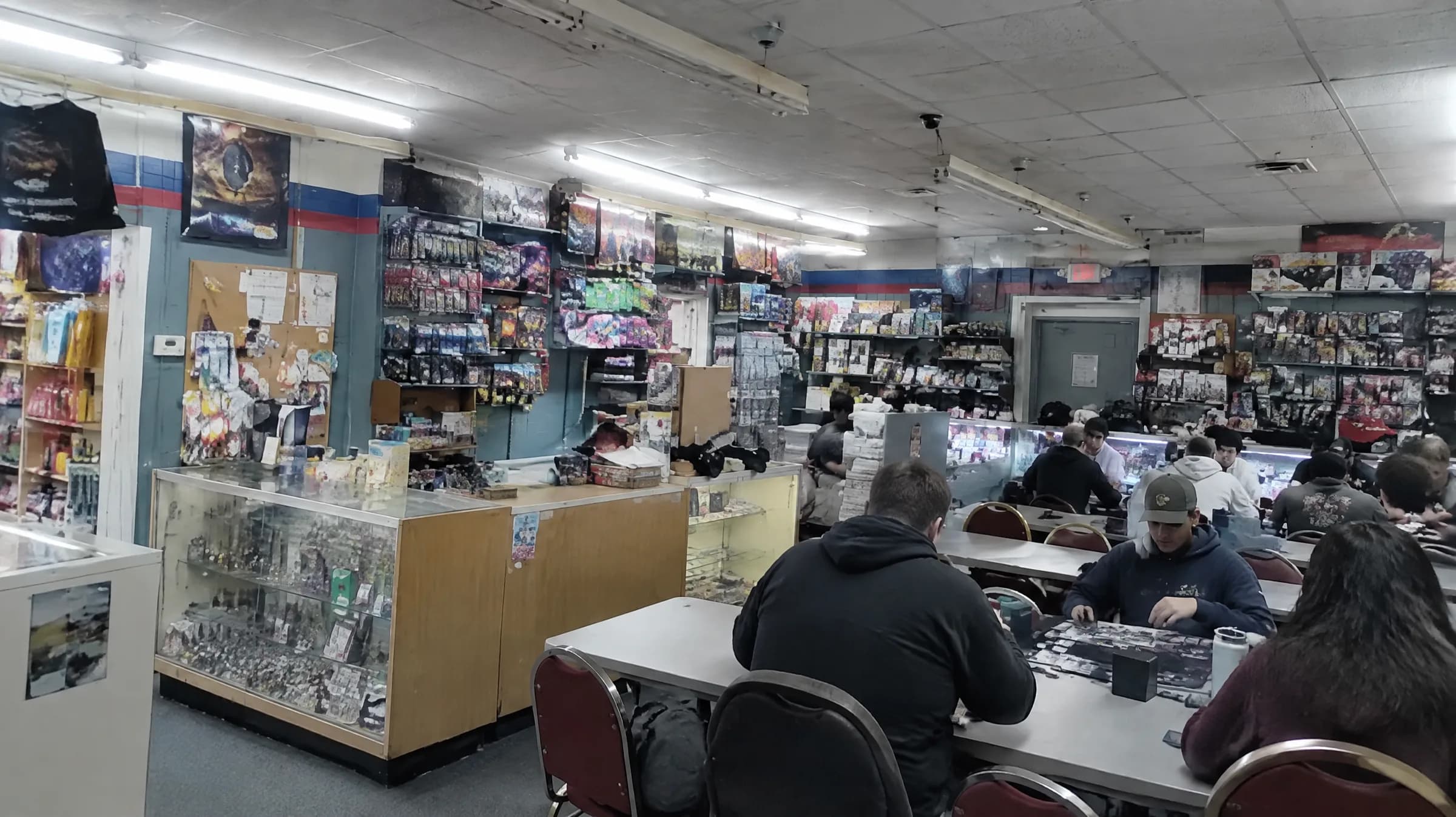 Inside Flights 2: glass cases of cards in front, wooden shelves of games, and players at folding tables under fluorescent lights.